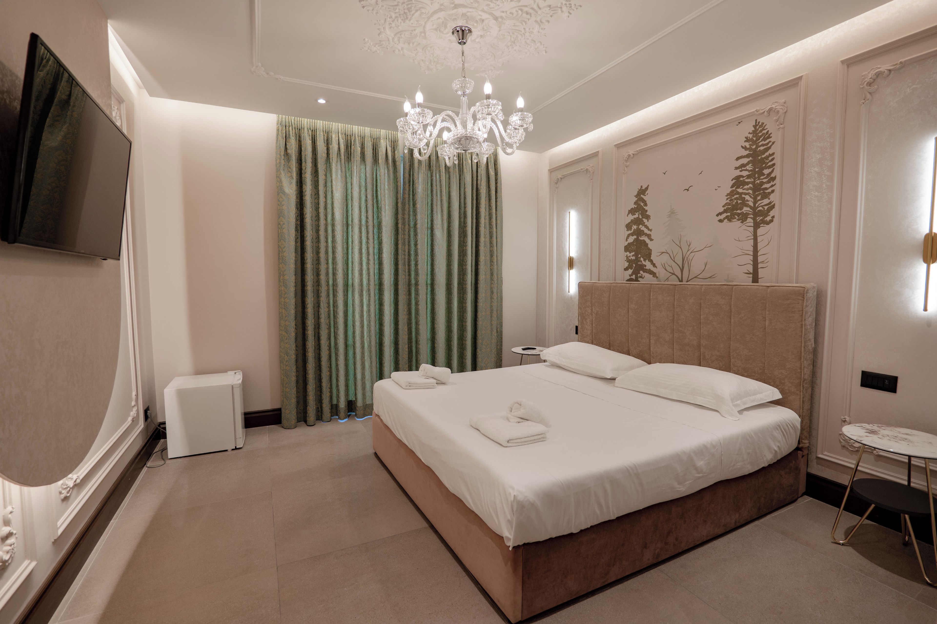 A modern hotel room with a large bed, a flat-screen TV, a chandelier, and a wall-mounted artwork of trees.