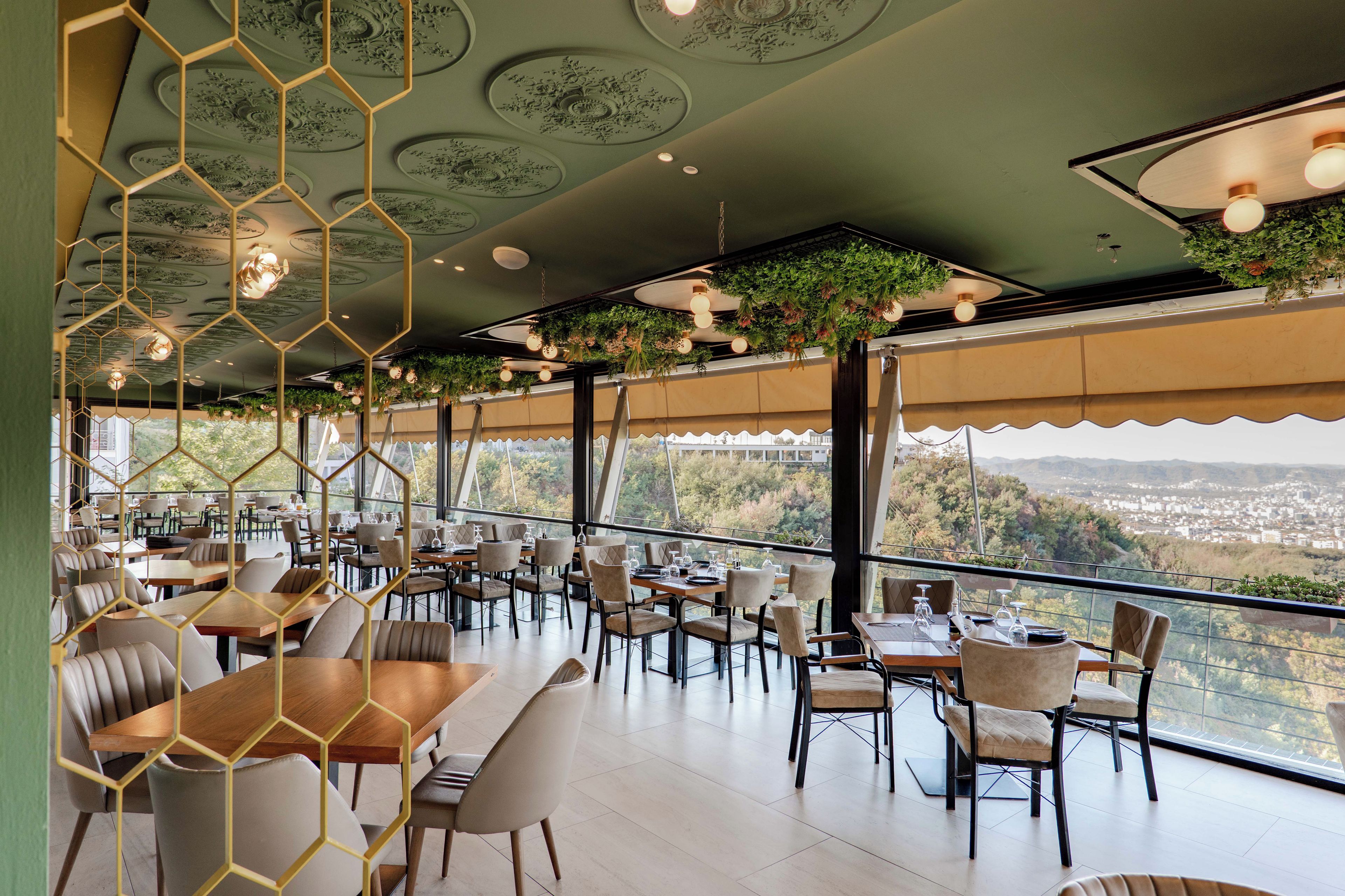A restaurant with a terrace overlooking a cityscape, featuring hexagonal ceiling decorations and hanging plants.