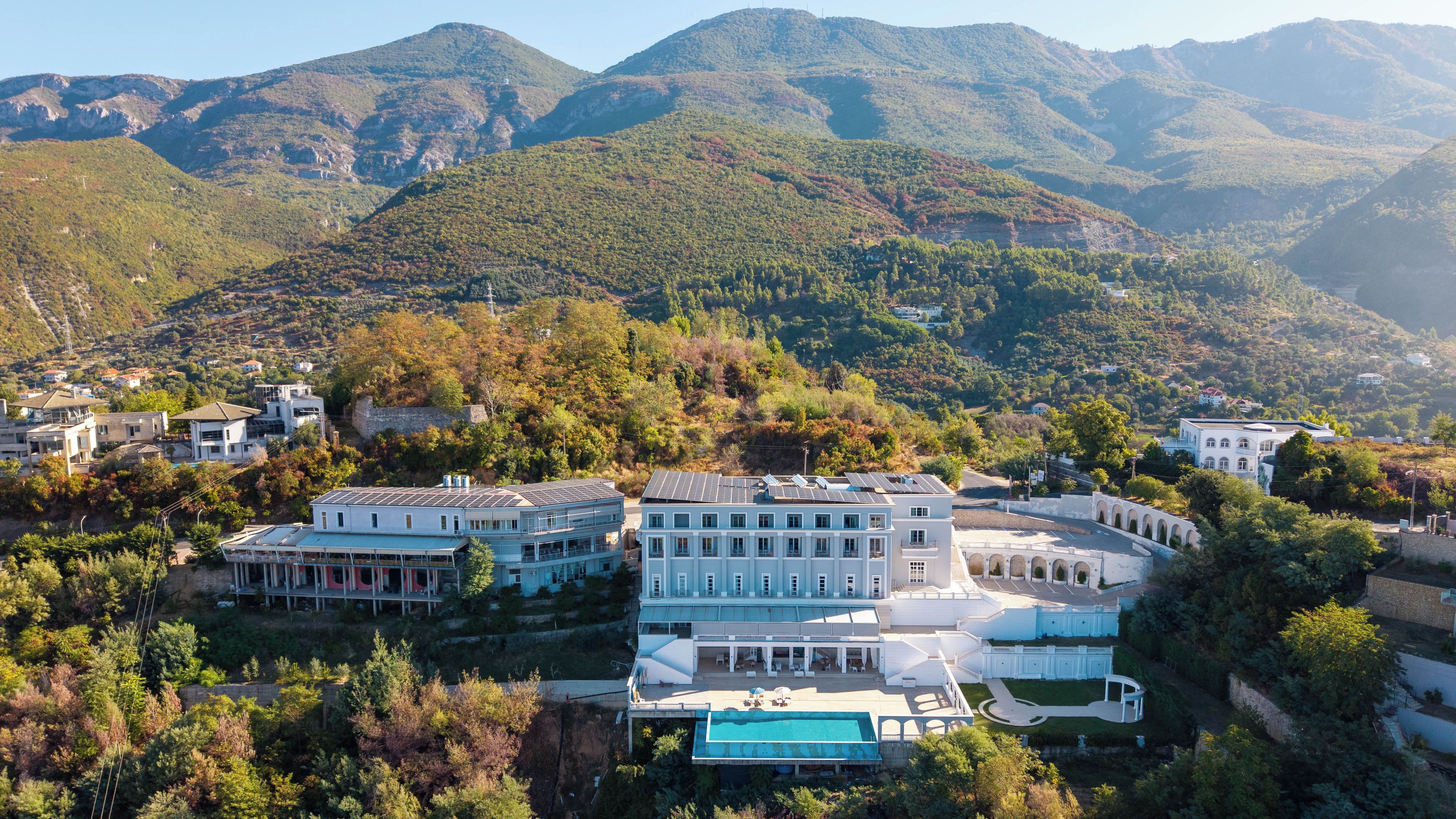 Un edificio grande de varios pisos con piscina, rodeado de abundante vegetación y montañas.