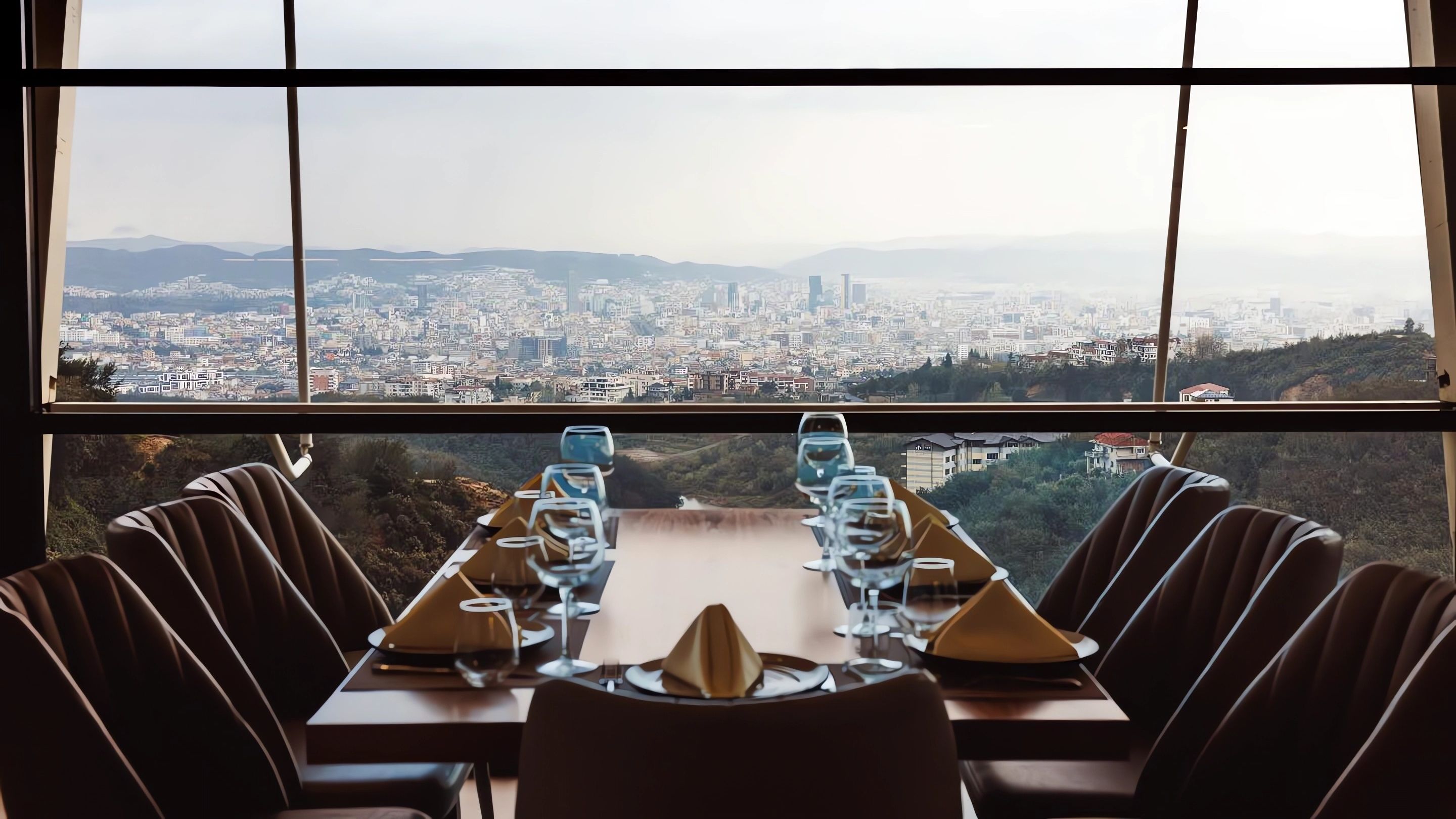 Une table à manger dressée pour six personnes, avec une vue sur la ville à travers de grandes fenêtres.