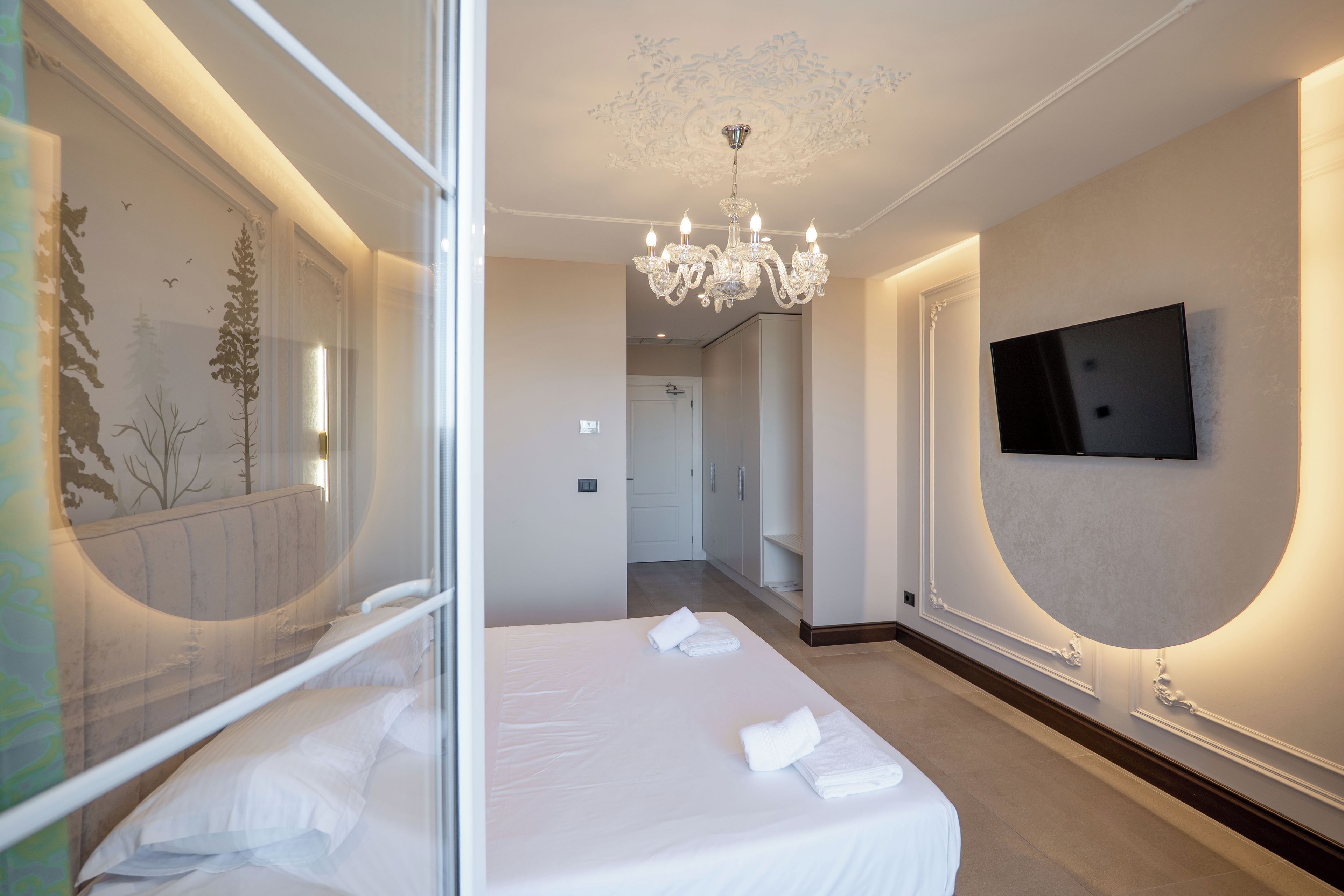A hotel room with a bed, a chandelier, a television mounted on the wall, and a hallway leading to another room.