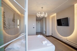 A hotel room with a bed, a chandelier, a television mounted on the wall, and a hallway leading to another room.