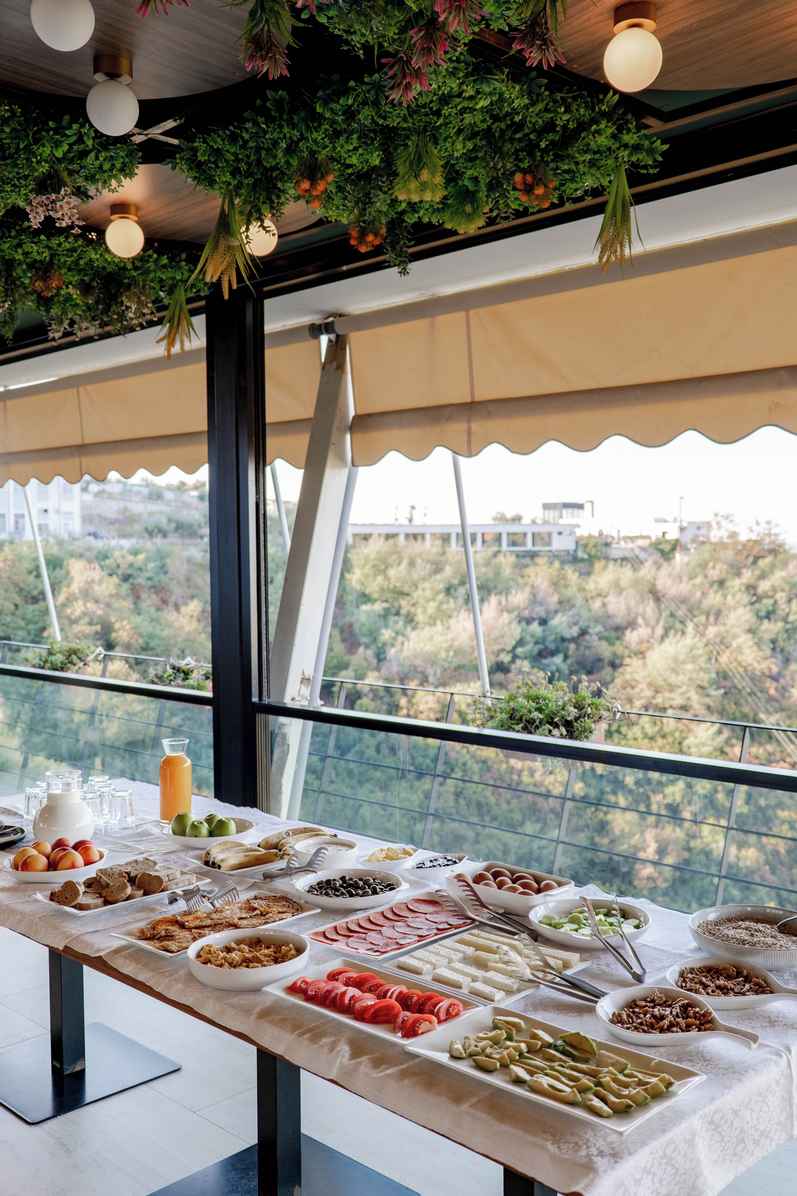 Una mesa de buffet con diversas opciones de comida y vista a un paisaje boscoso a través de amplias ventanas.