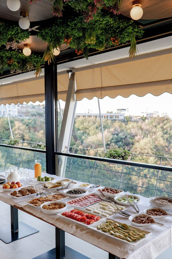 A buffet table with a variety of food items and a view of a forested landscape through large windows.