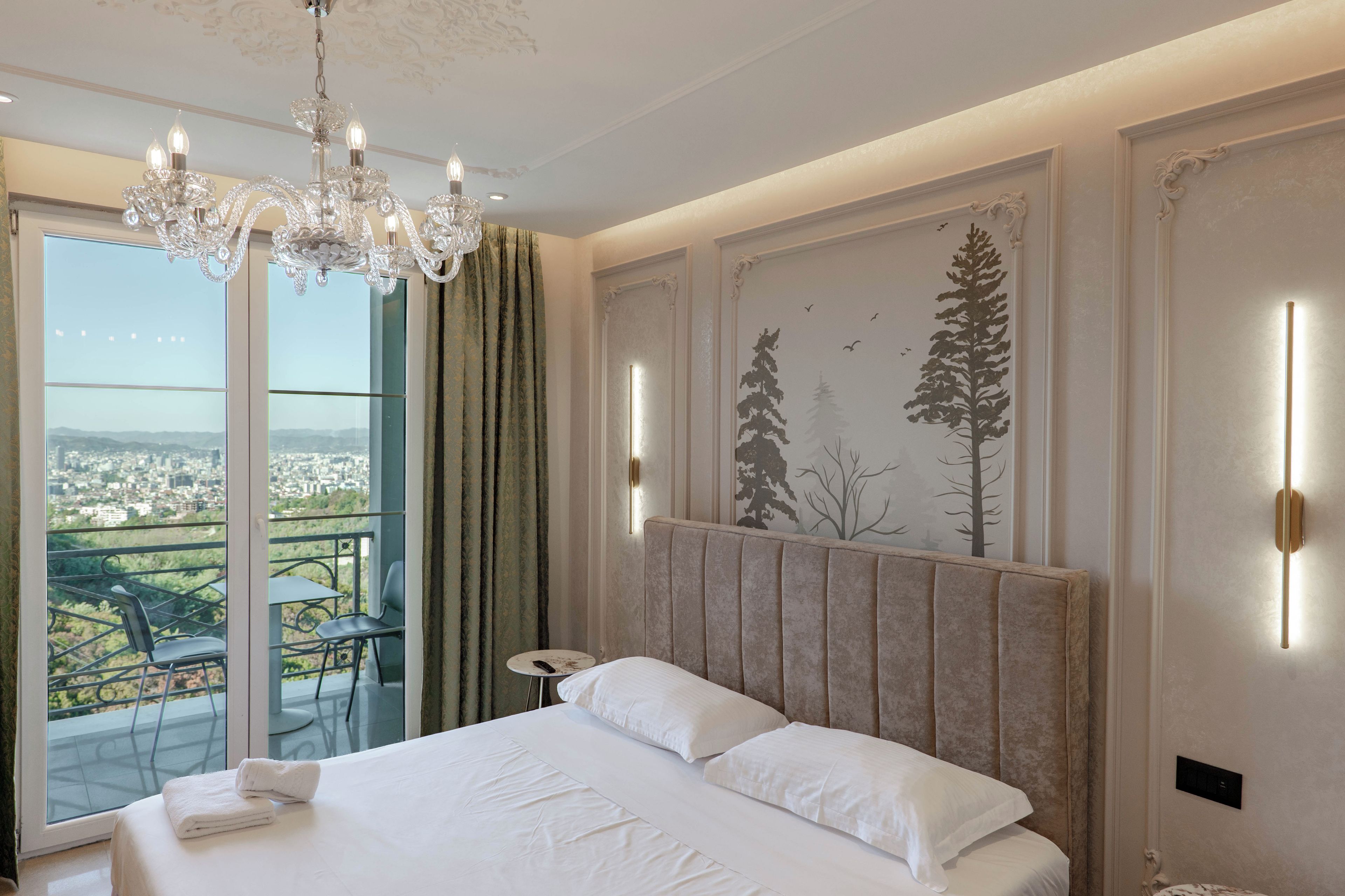Habitación de hotel con una cama grande, un candelabro, un balcón con mesa y sillas, y vista a la ciudad.