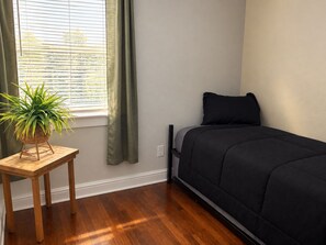 A bedroom with a bed, a small table with a plant, and a window with blinds.