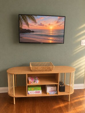 Un televisor montado en una pared muestra una puesta de sol sobre el océano. Abajo, una consola de madera sostiene una cesta tejida y juguetes infantiles.