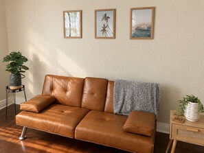 Un sofá de cuero marrón con una manta, una mesita de noche con una planta y cuadros enmarcados en la pared.
