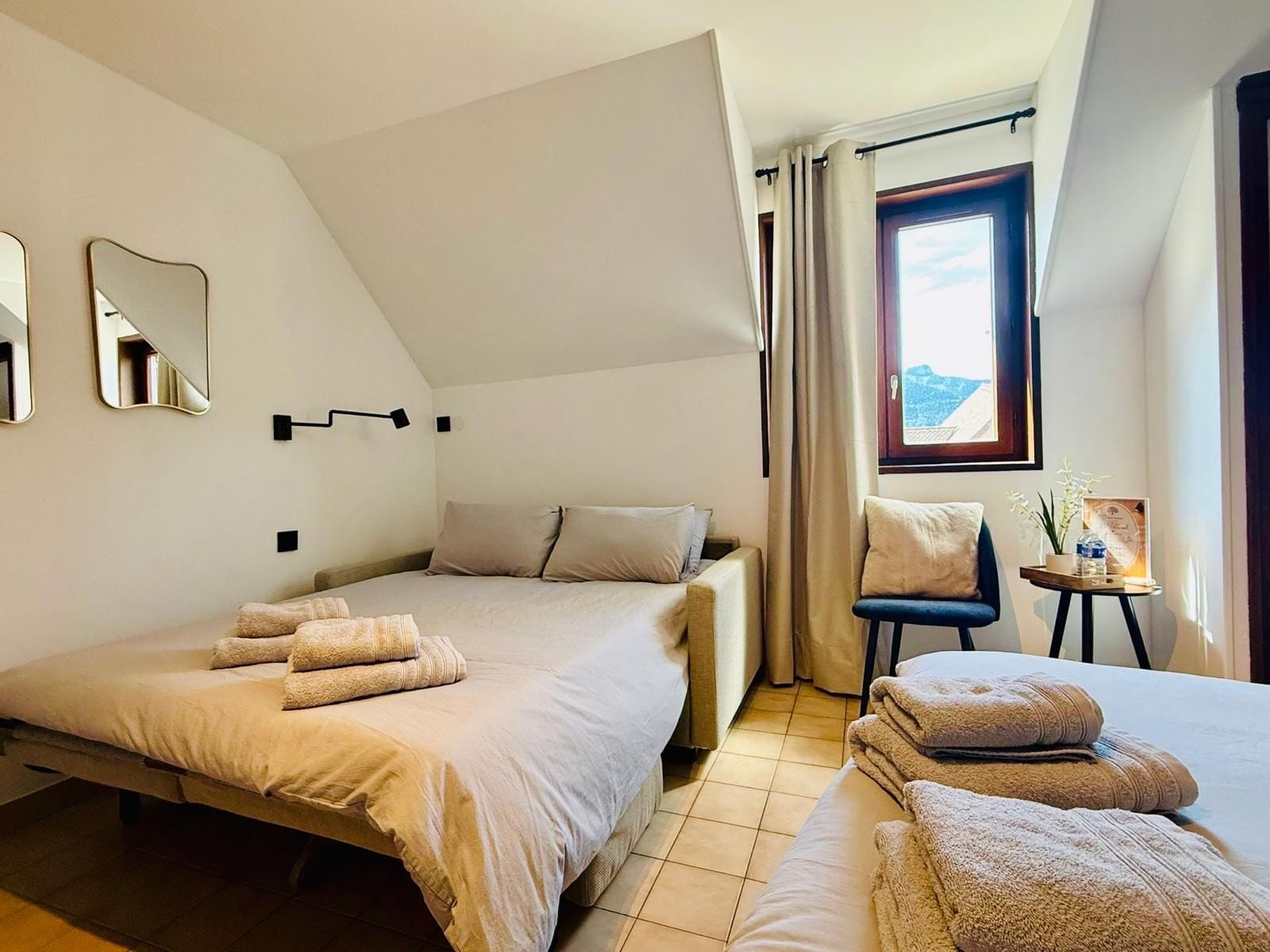 A modern hotel room with a bed, a chair, a small table, and a window with curtains.