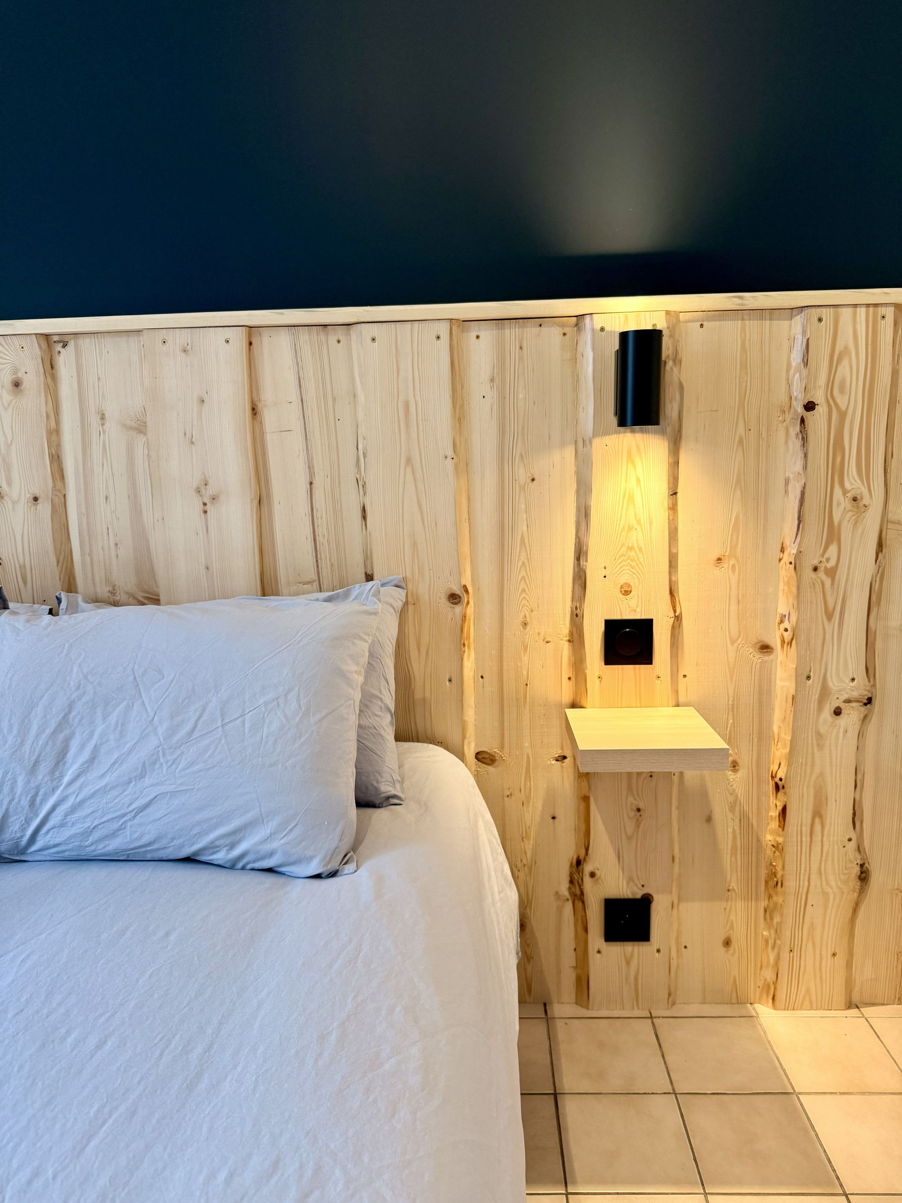 Un dormitorio con cabecera de madera, cama con ropa de cama blanca, un estante pequeño y un aplique en la pared.