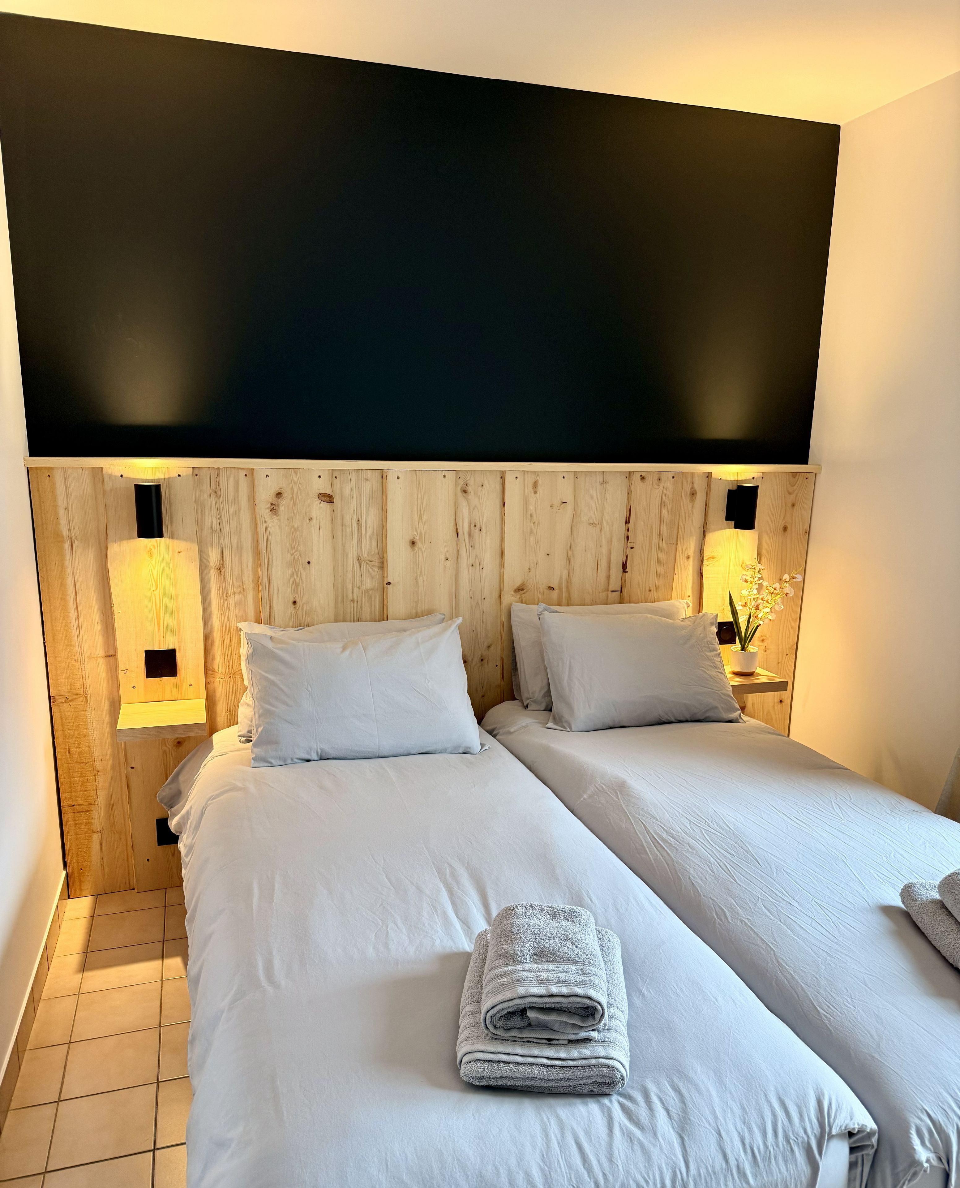 Quarto de hotel com cabeceira de madeira, duas camas, roupa de cama branca, toalha dobrada e luminárias de cabeceira.
