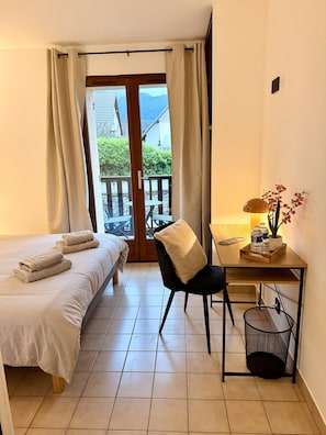 A hotel room with a bed, a desk with a chair, a small table with a lamp and flowers, and a view of the outdoors through a glass door.