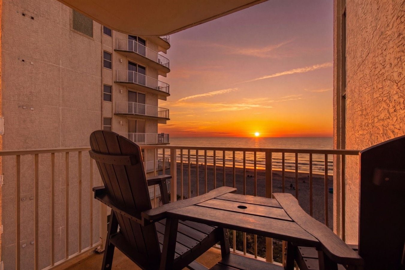 Un balcon avec une table et des chaises en bois, d’où l’on peut admirer le coucher de soleil sur l’océan.