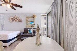 A hotel room with a bed, a sofa, a dining table, a decorative wall piece, a ceiling fan, and a view through a window. - Long Beach Resort Beachfront Studio with Ocean View Balcony, Sleeps 4, Tower 1 (Panama City Beach)