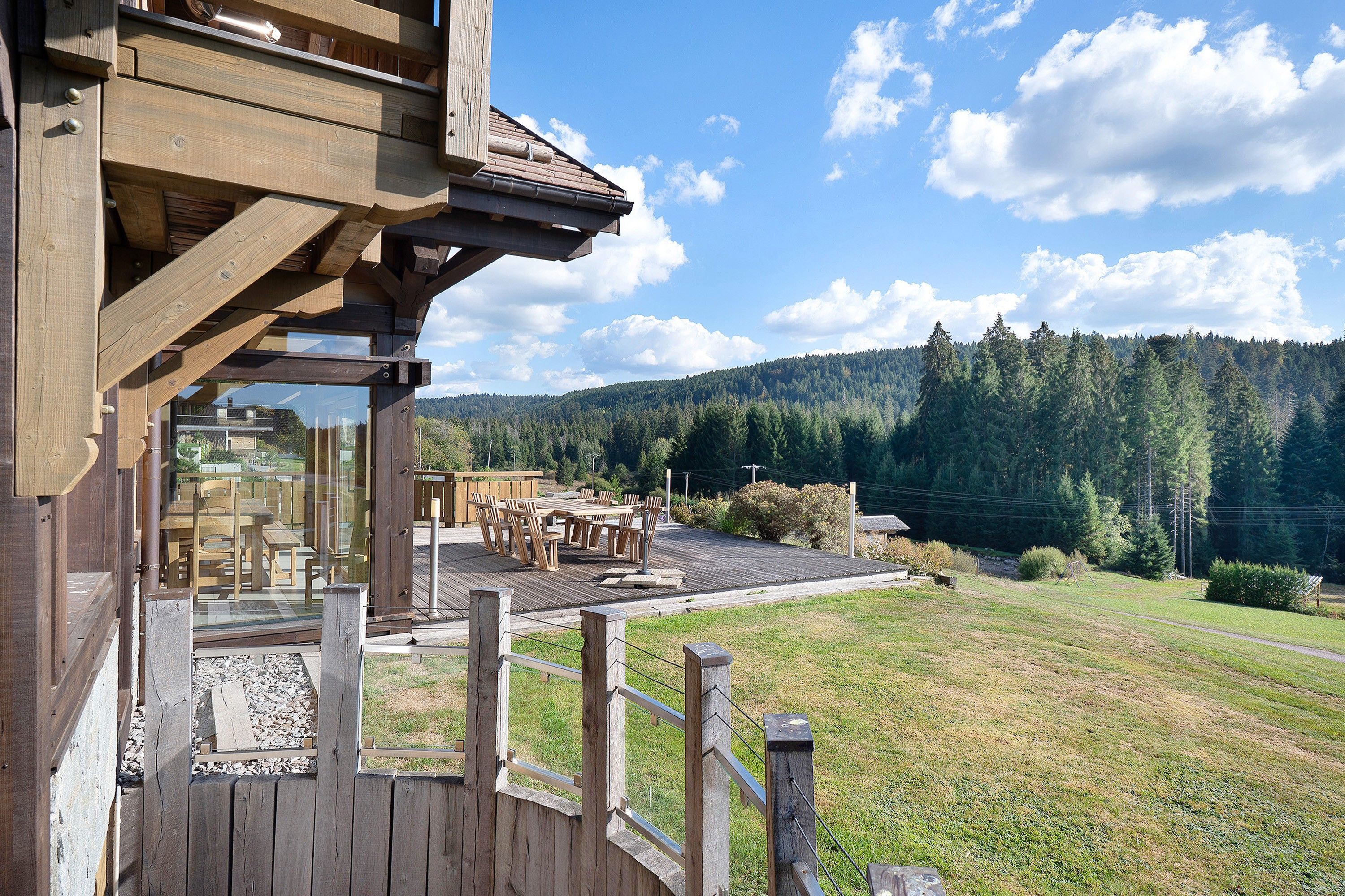A wooden building with a deck and outdoor seating area, overlooking a forested hillside.