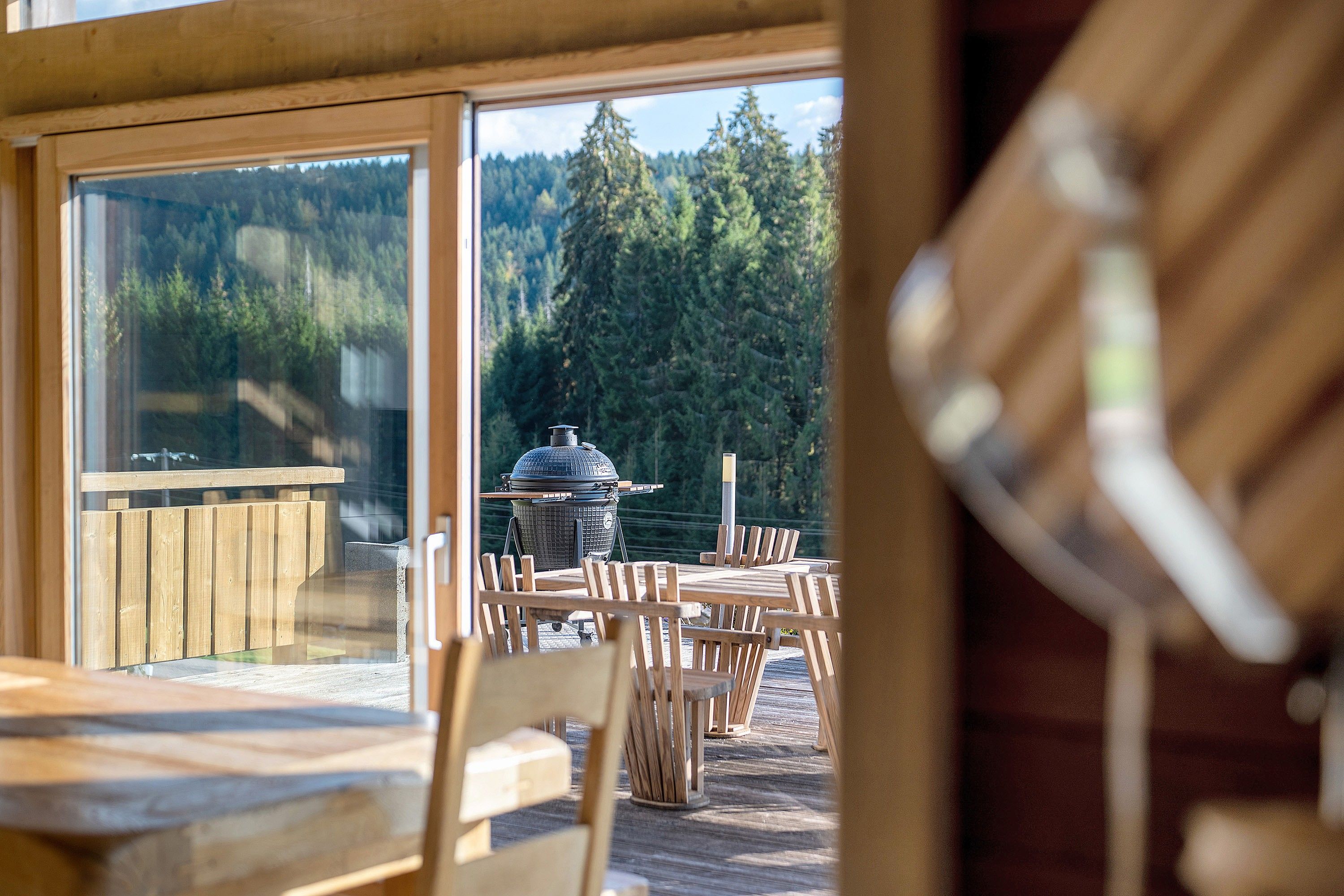 An outdoor dining area with wooden tables and chairs, a barbecue grill, and a view of a forest through large windows.