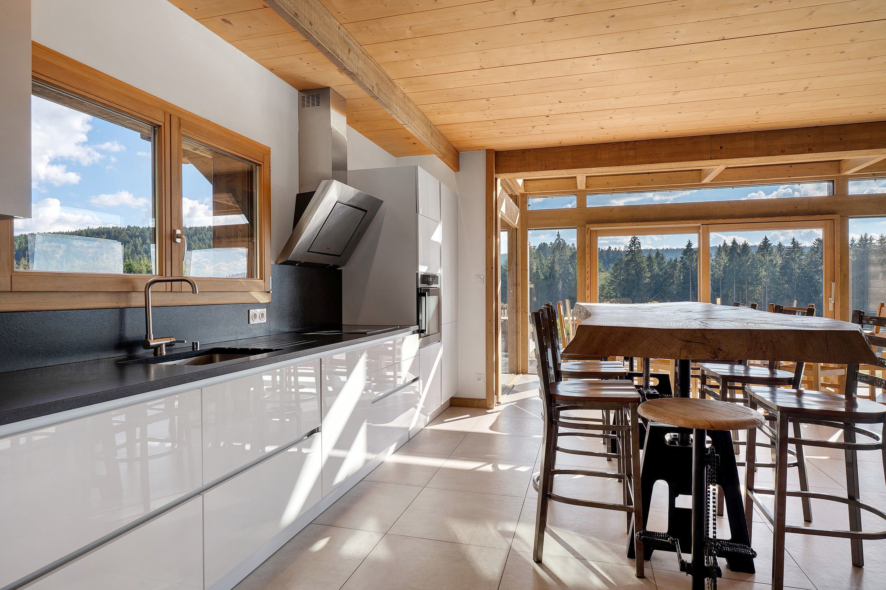 A modern kitchen with a large island, a dining area with a wooden table and chairs, and large windows offering a view of a forested landscape.