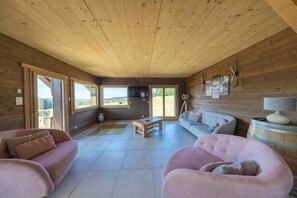 A spacious living room with a wooden ceiling, large windows, a sofa, a coffee table, and a TV. - Chalet 'L'Alpage' with Mountain View, Sauna and Jacuzzi (Gérardmer)