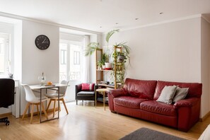 Una sala de estar con un sofá de cuero rojo, una mesa de comedor con sillas, un reloj en la pared y una estantería con plantas.