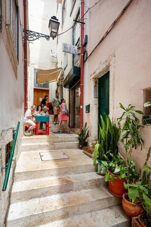 A narrow street with steps leading to a building entrance, potted plants, and a table with people sitting. - Alfama 2BR near Sé & Terreiro do Paço (Lisbon)