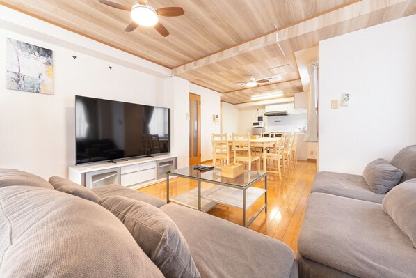 A modern living room with a flat-screen TV, a glass coffee table, and a dining area with wooden chairs.