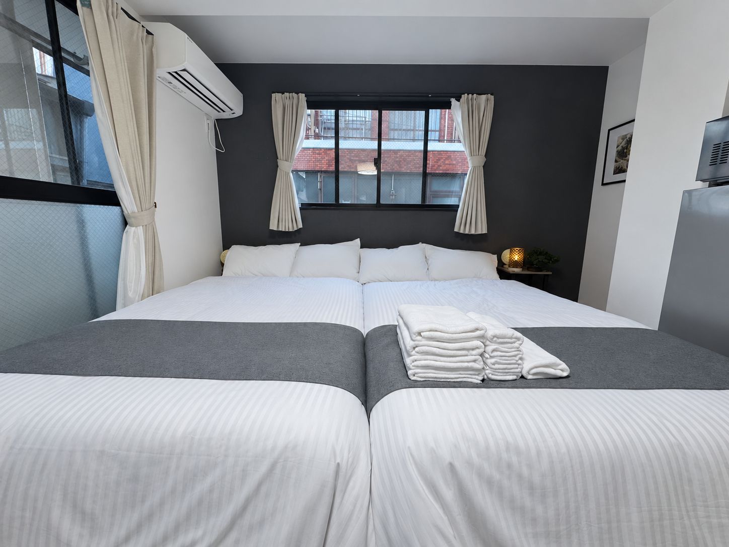 Ein Hotelzimmer mit einem Bett, zwei Nachttischen, einem Fenster mit Vorhängen, einer Klimaanlage und einer Wandlampe.
