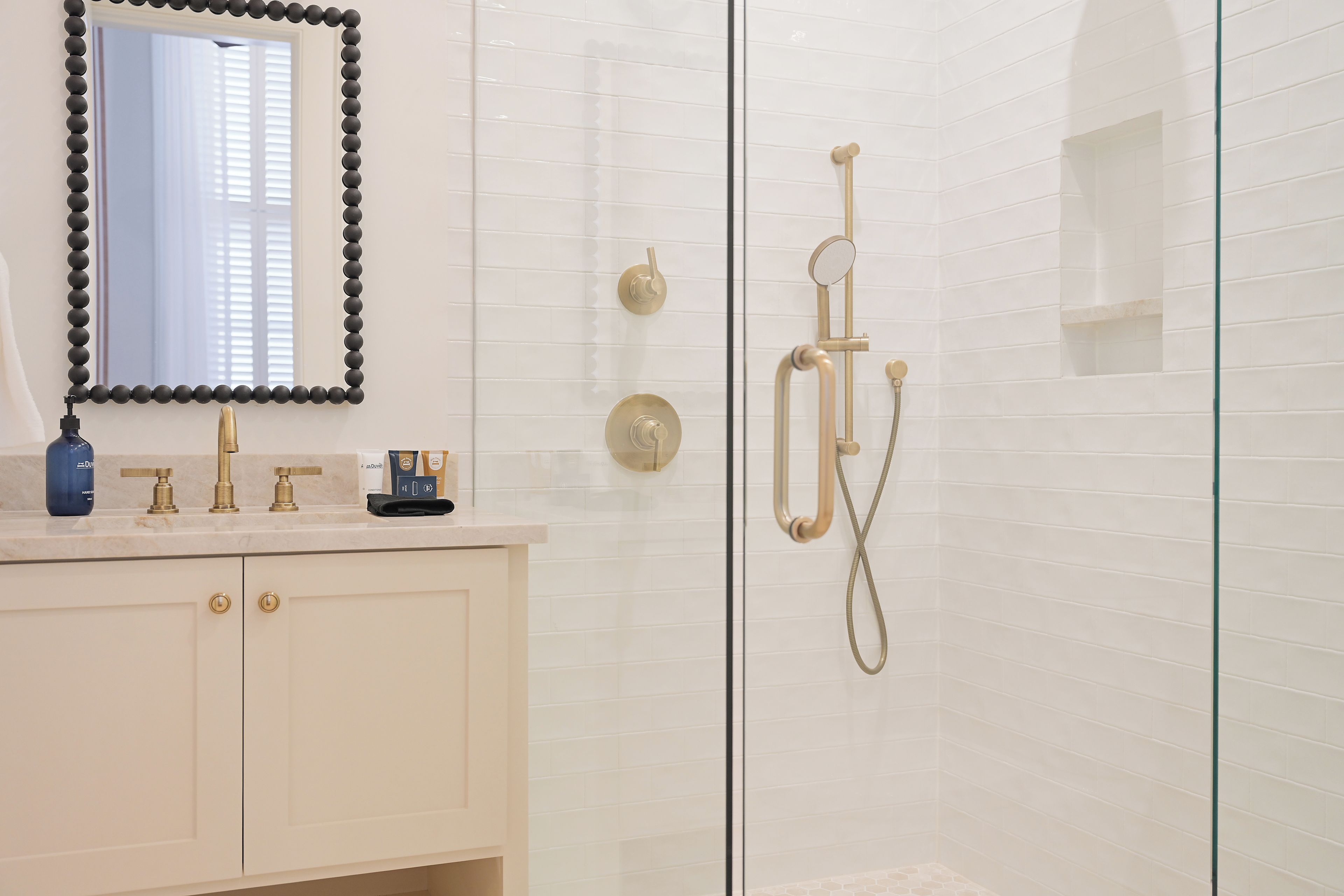 Un baño con cabina de ducha revestida de azulejos blancos, mampara de vidrio y tocador con espejo y dispensador de jabón.