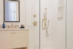 Un baño con cabina de ducha revestida de azulejos blancos, mampara de vidrio y tocador con espejo y dispensador de jabón.