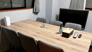 Sala de conferência com uma mesa comprida, cadeiras, uma TV e um orador.