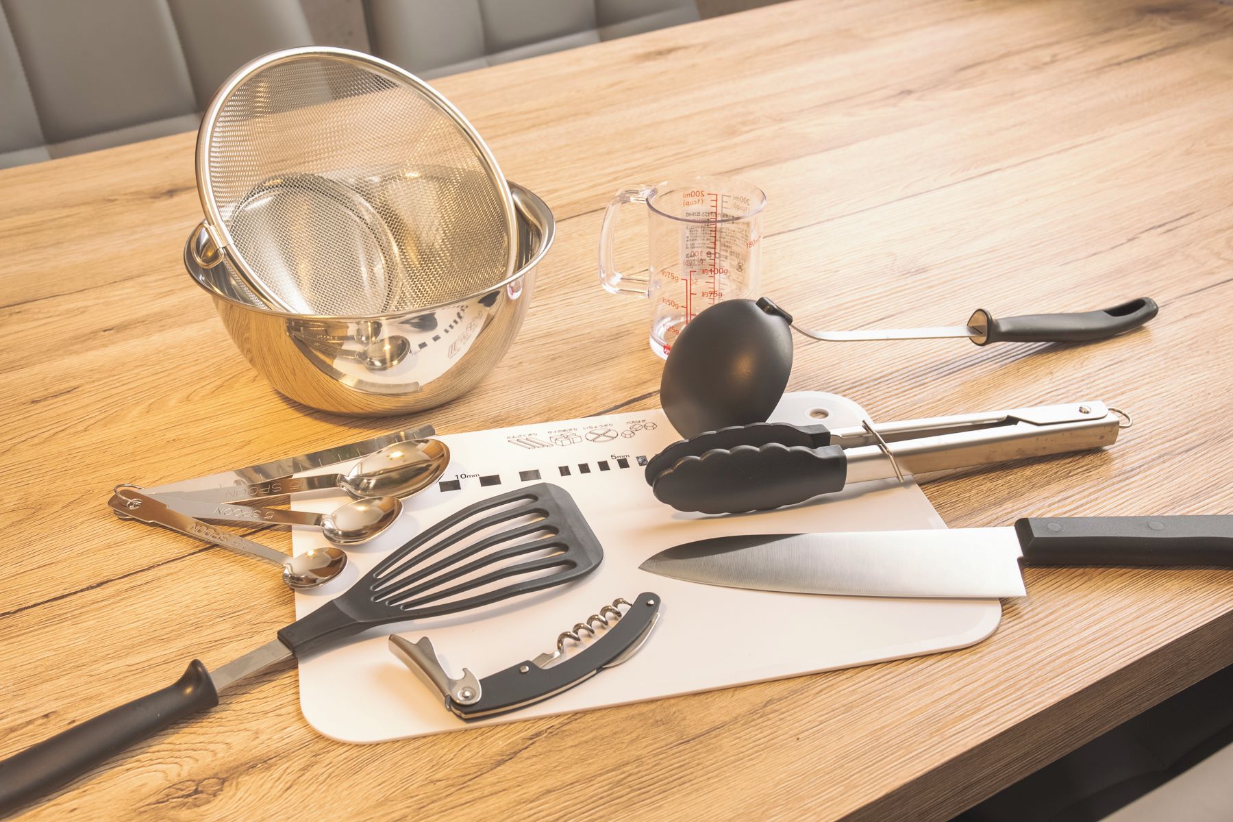 Uma mesa de madeira com utensílios de cozinha, incluindo um escorredor, medidor, espátula, pinças e uma faca.