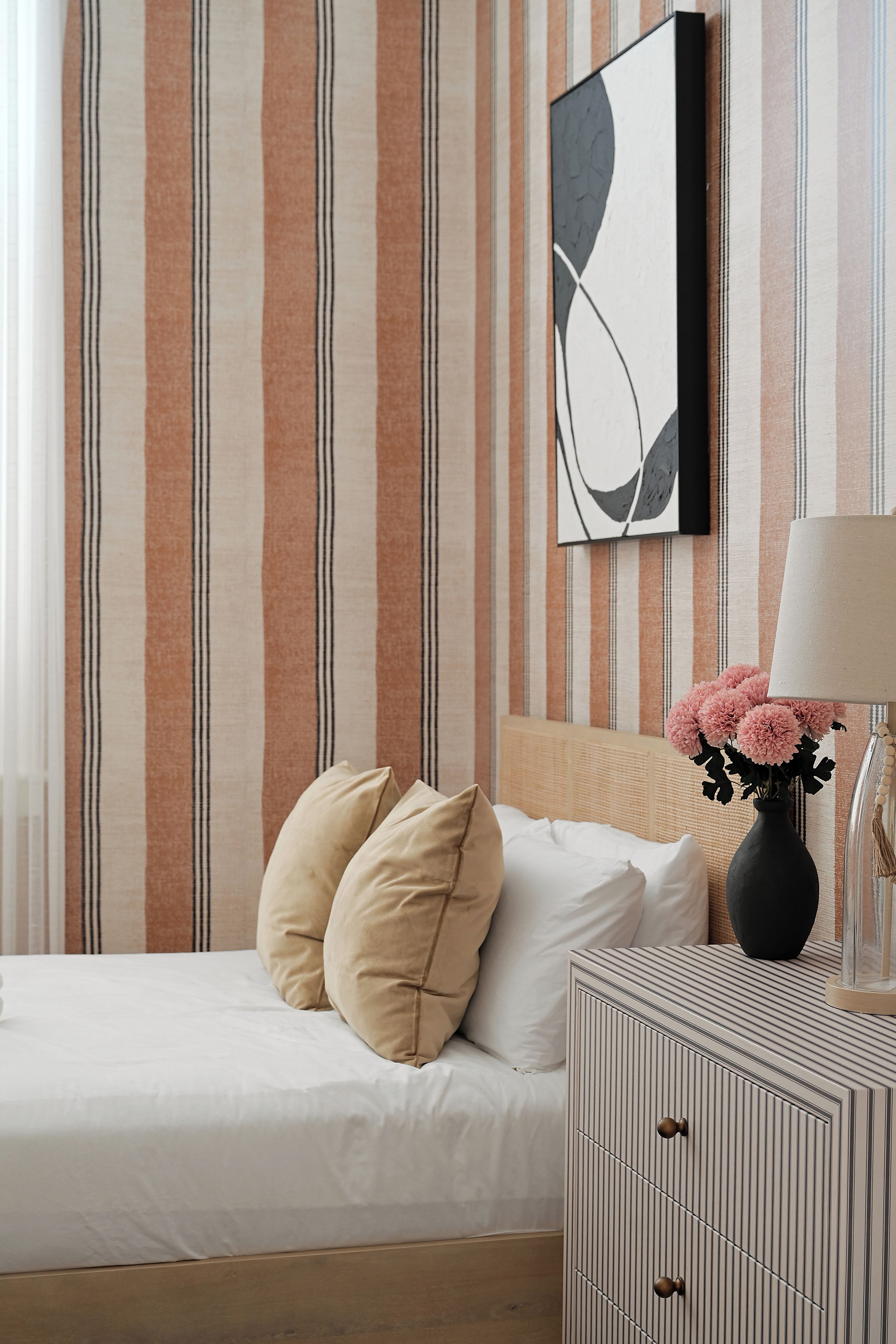 Una cama bien hecha con dos almohadas, una mesita de noche con un florero y una pared a rayas con una obra de arte abstracta.