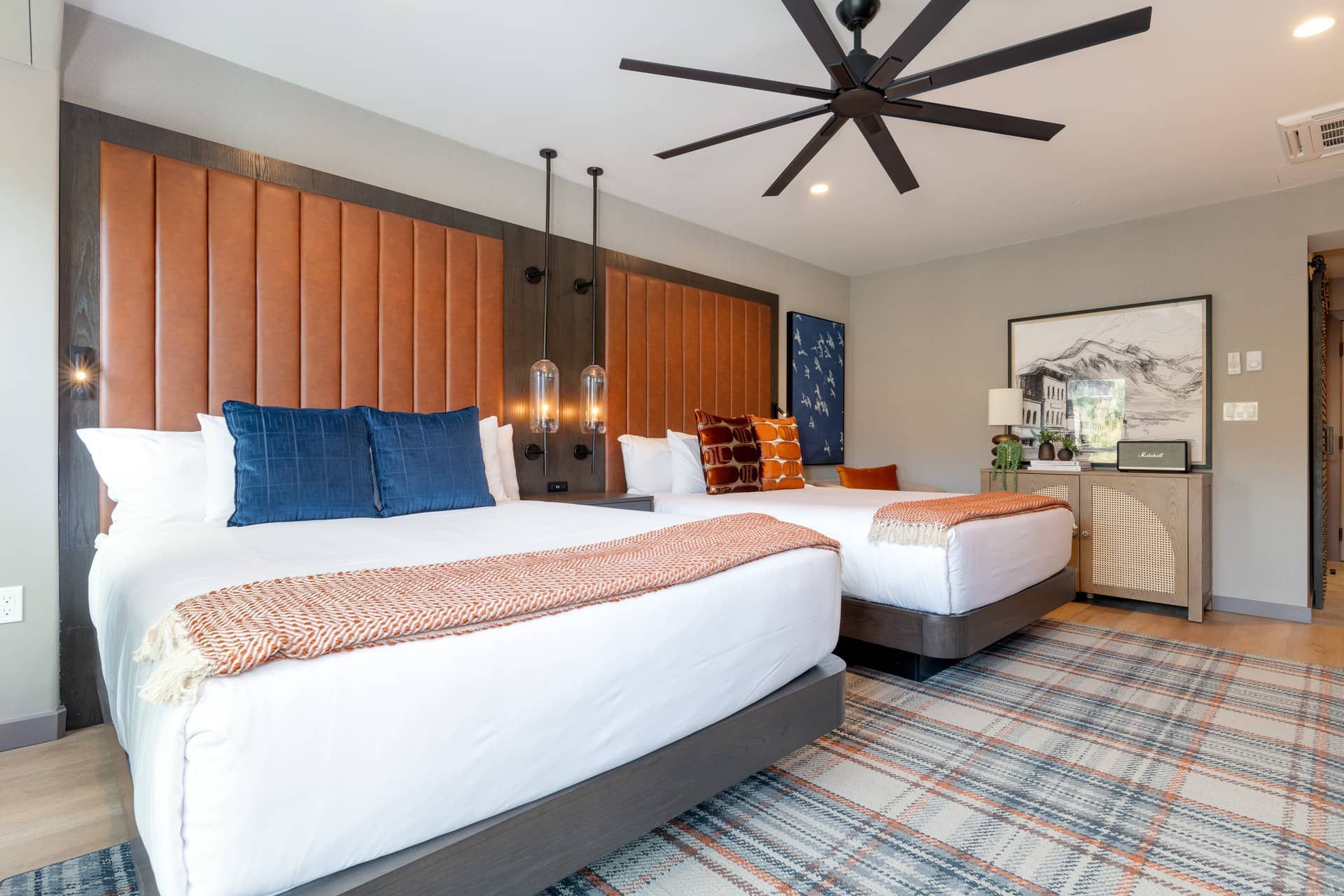 A hotel room with two beds, a ceiling fan, a wall-mounted light fixture, and a framed picture on the wall.