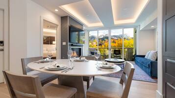 A modern dining area with a round table, chairs, and a view of autumn foliage through large windows.