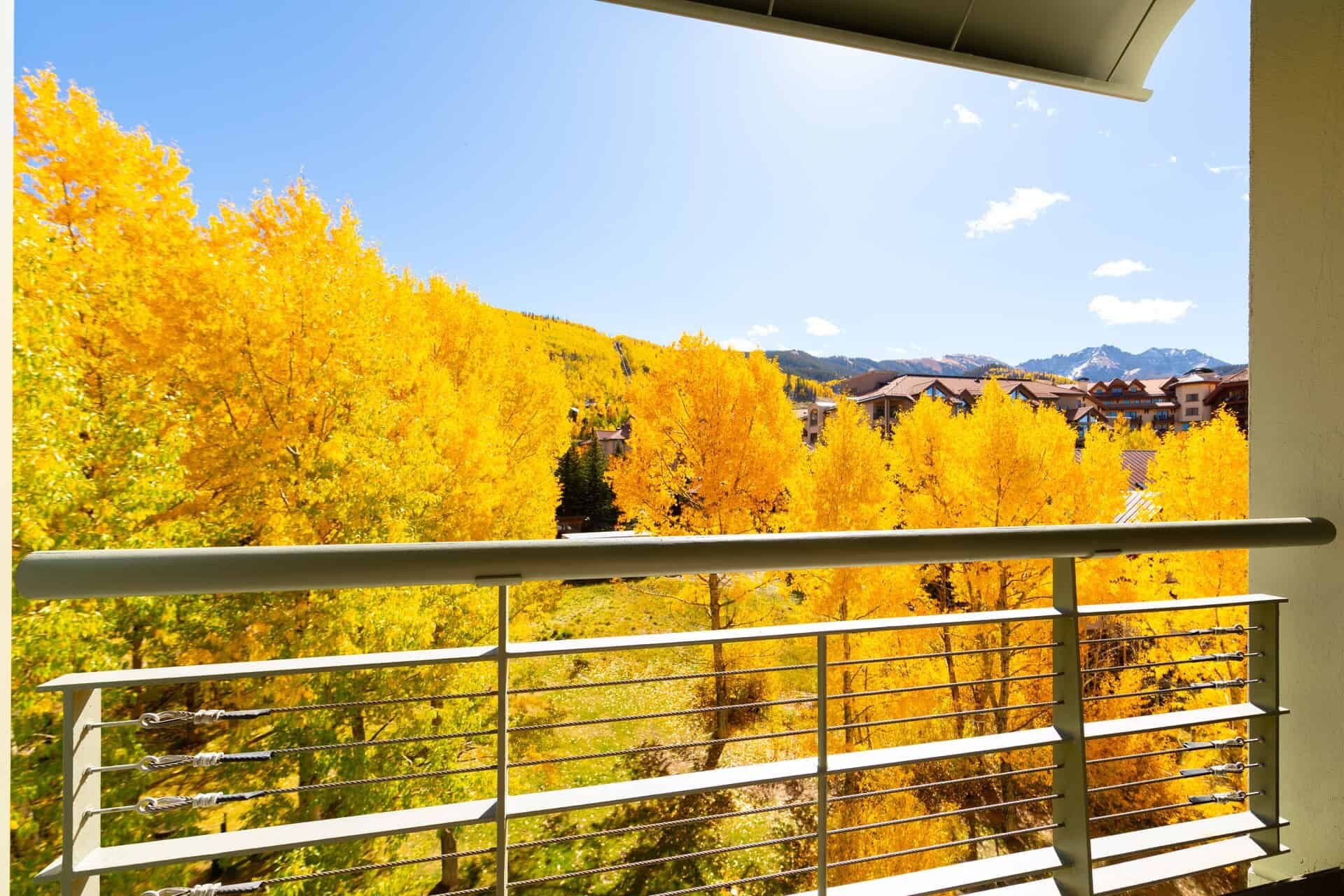 A balcony with a metal railing overlooking a mountainous area with autumn-colored trees and a clear sky.