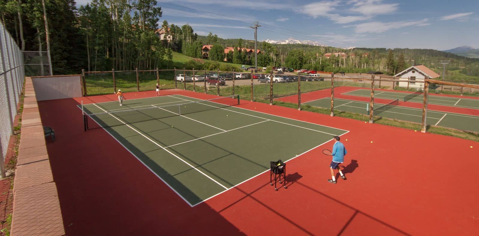 An outdoor tennis court with multiple courts, surrounded by a fence, and a parking area in the background.