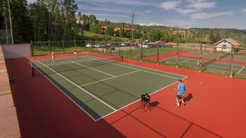 An outdoor tennis court with multiple courts, surrounded by a fence, and a parking area in the background.