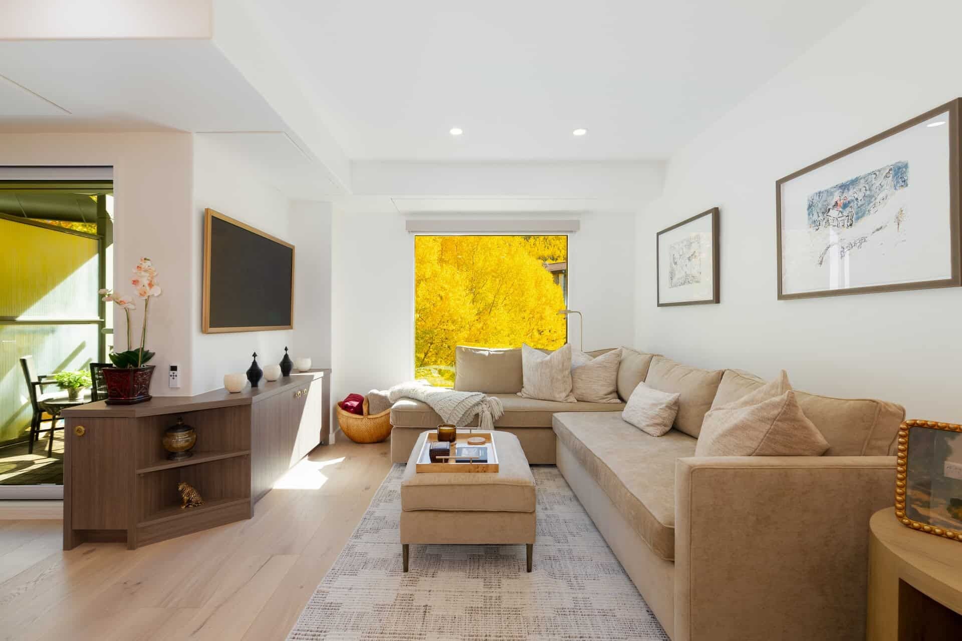 Un salon moderne avec un canapé, un pouf et une vue sur le feuillage jaune à travers une fenêtre.