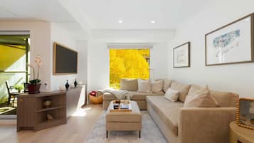 A modern living room with a sofa, ottoman, and a view of yellow foliage through a window.