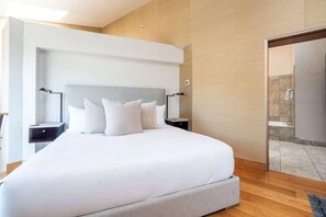 Ein modernes Hotelzimmer mit großem Bett, Nachttischen, einer Ablage und einer begehbaren Garderobe sowie einem Duschbereich.