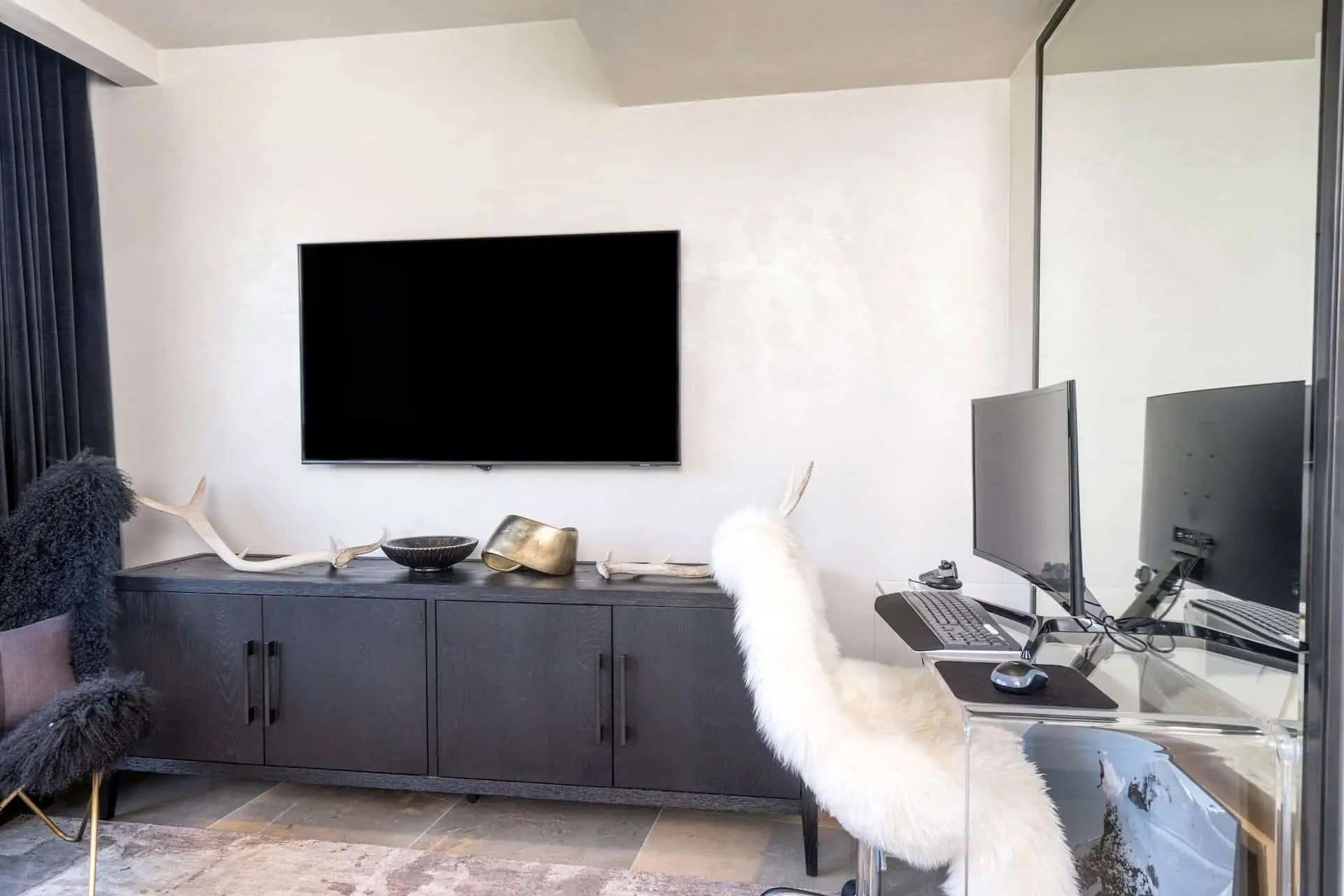 Una sala de estar moderna con un televisor de pantalla plana montado en la pared, un mueble de madera oscura, un escritorio de vidrio con una computadora y una silla blanca mullida.