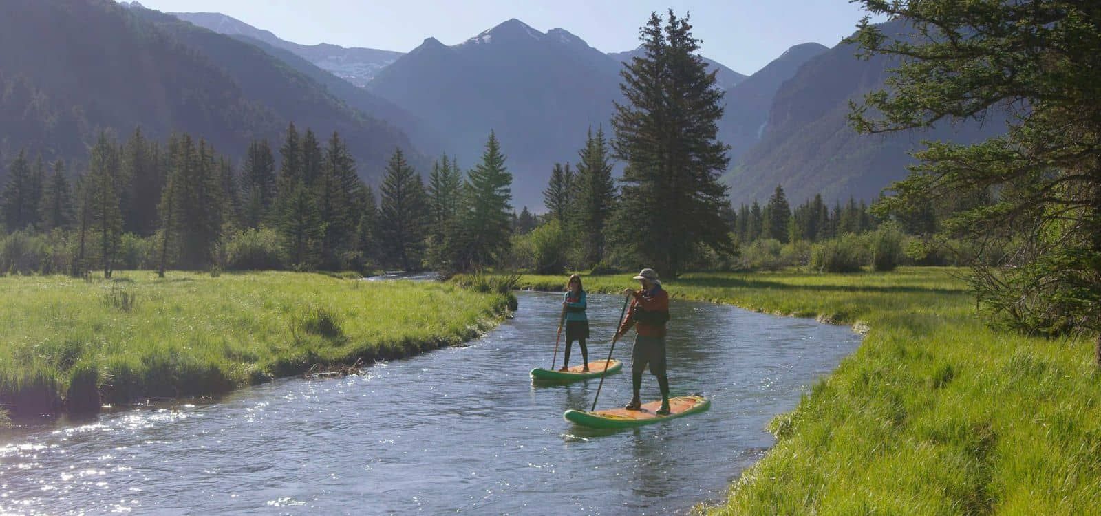 Zwei Personen stehen auf Paddleboards auf einem Fluss, umgeben von Bergen und viel Grün.