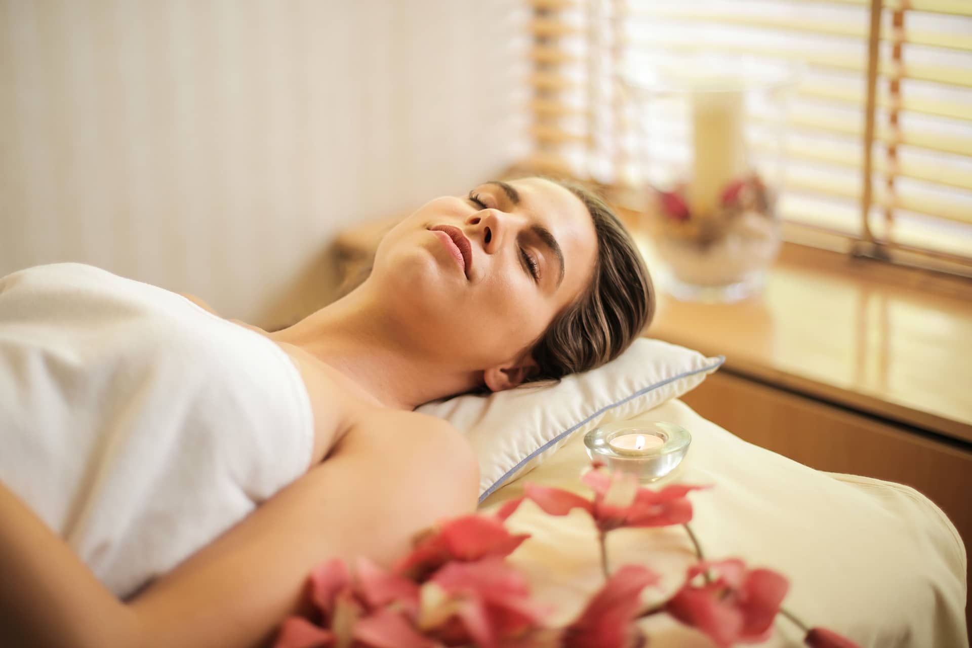 A person lying on a bed with a pillow and a small table with a candle and flowers beside them.