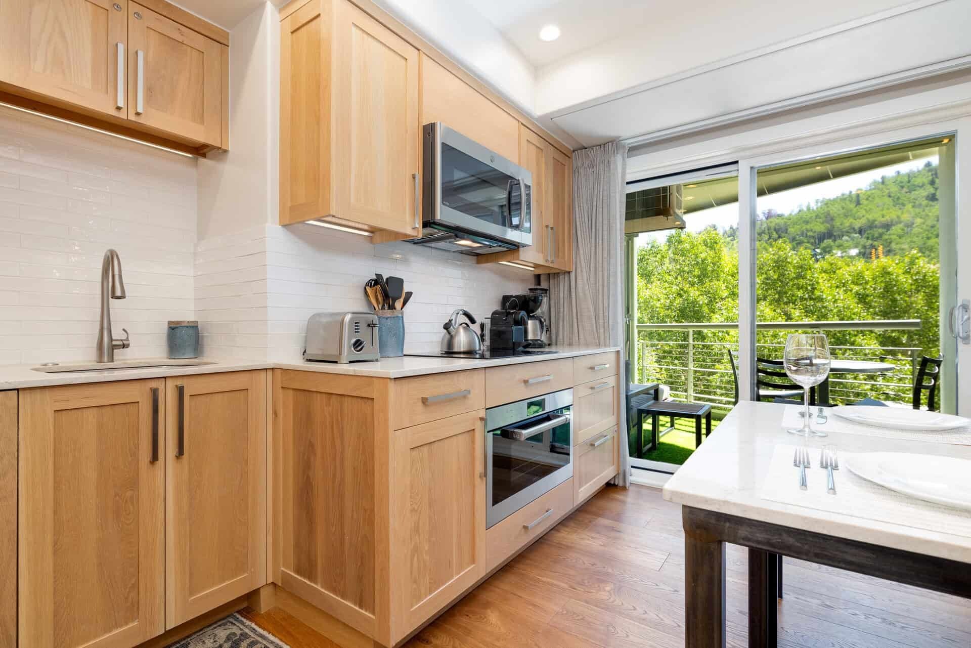 A modern kitchen with wooden cabinets, a microwave, and a toaster, leading to a balcony with a table and chairs.