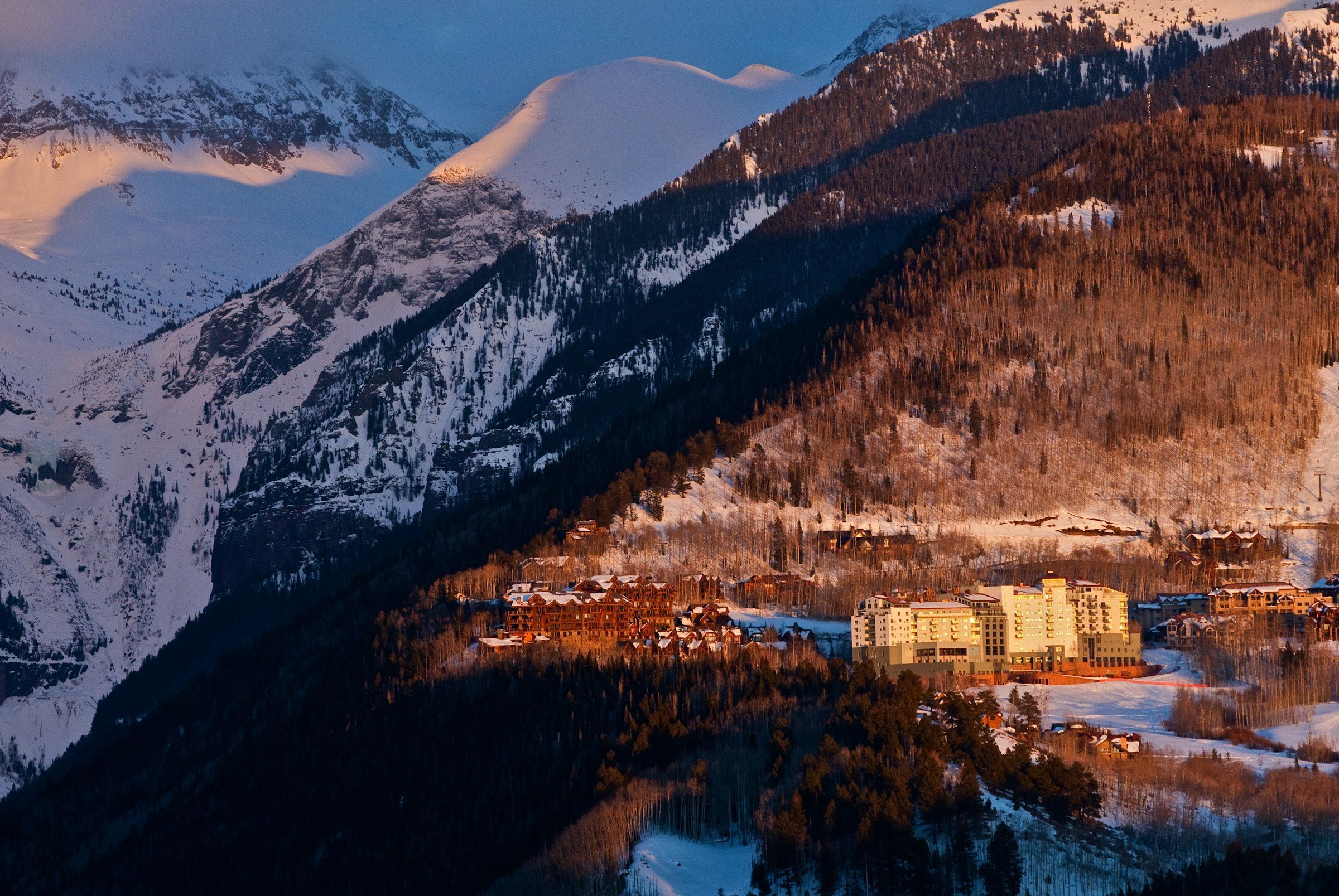 A mountain resort with a large lodge and cabins, surrounded by snow-covered peaks and forested slopes.