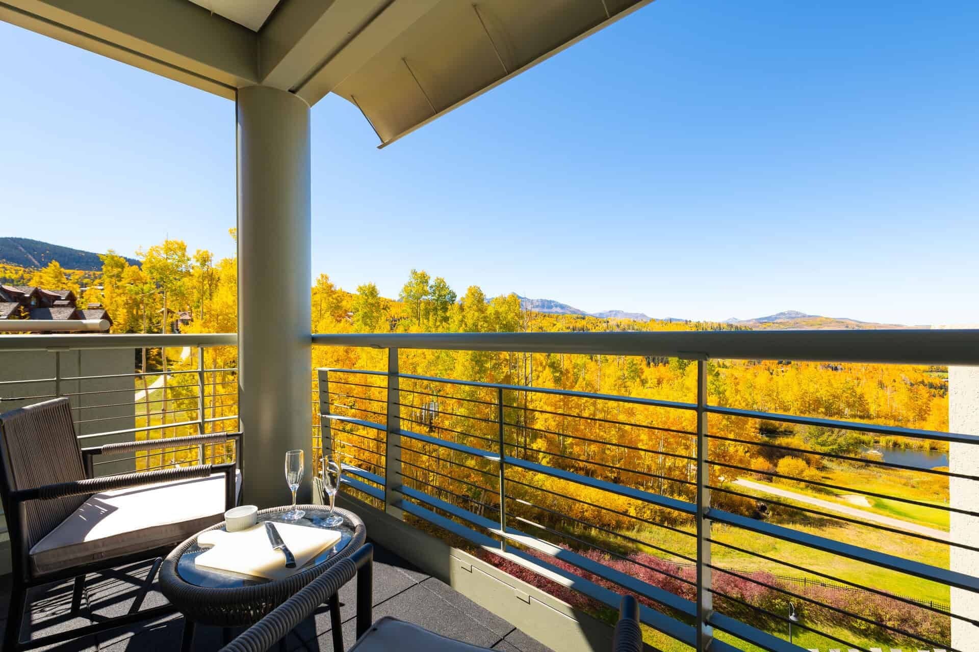 A balcony with a table and chairs, overlooking a landscape with autumn-colored trees and distant mountains.