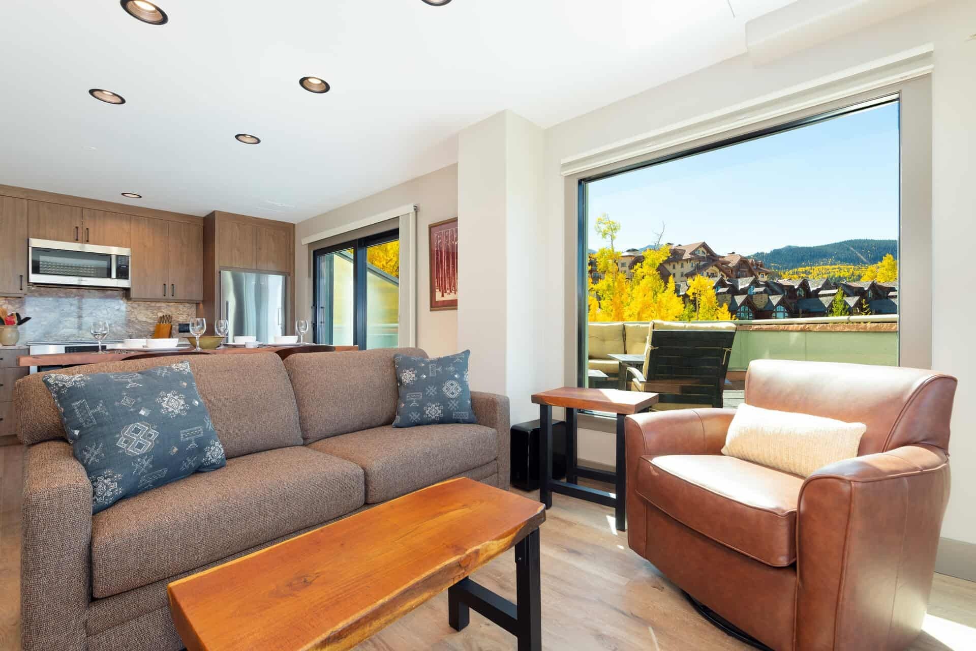 A modern living room with a sofa, a chair, and a coffee table. There is a kitchen area with a microwave and cabinets. A large window offers a view of a vineyard and buildings.