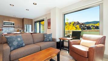 A modern living room with a sofa, a chair, and a coffee table. There is a kitchen area with a microwave and cabinets. A large window offers a view of a vineyard and buildings.