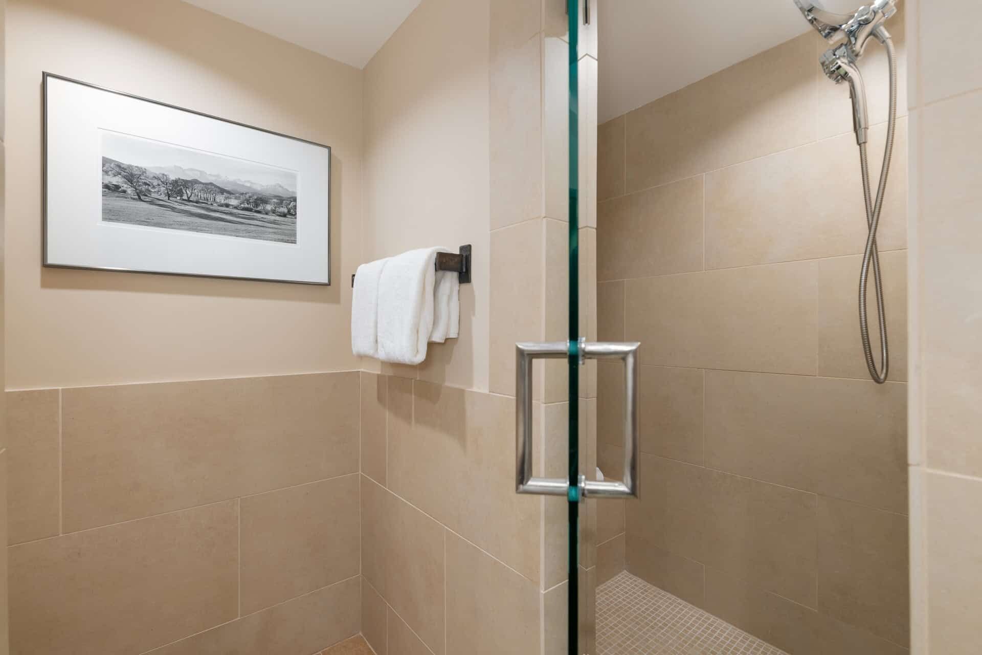 Una ducha con cerramiento de vidrio, ducha de mano, paredes con azulejos color beige y una fotografía blanco y negro enmarcada en la pared.