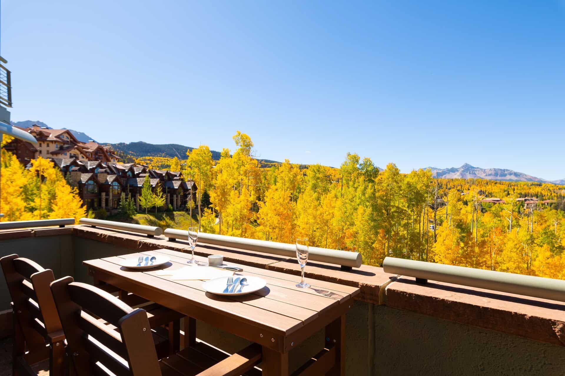 An outdoor dining area with a table set for two, overlooking a forest with autumn foliage and mountains in the background.