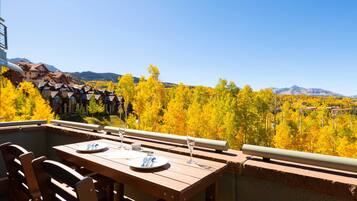 An outdoor dining area with a table set for two, overlooking a forest with autumn foliage and mountains in the background.